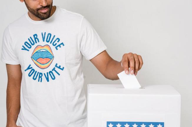 A man casts his vote into a ballot box, wearing a 'Your Voice Your Vote' t-shirt, promoting active participation in democracy.